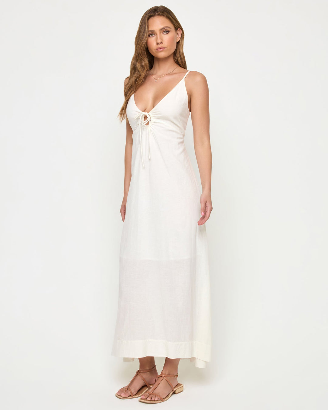 Three-quarter view of a model wearing the LSPACE Mabel Maxi Dress in cream, featuring thin spaghetti straps, a deep V-neckline with ruched drawstring tie at the bust, and a flowing full-length silhouette.