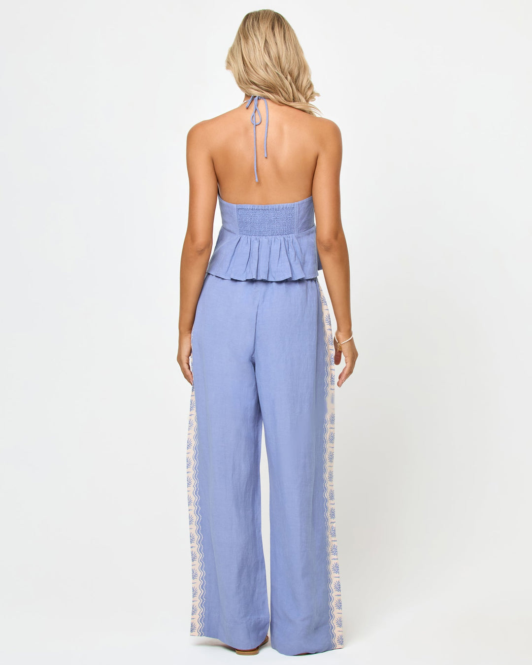 Back view of a model wearing the LSPACE April Top in shell, showing the open back with halter neck tie, smocked band, and peplum ruffle hem, paired with matching wide-leg pants.