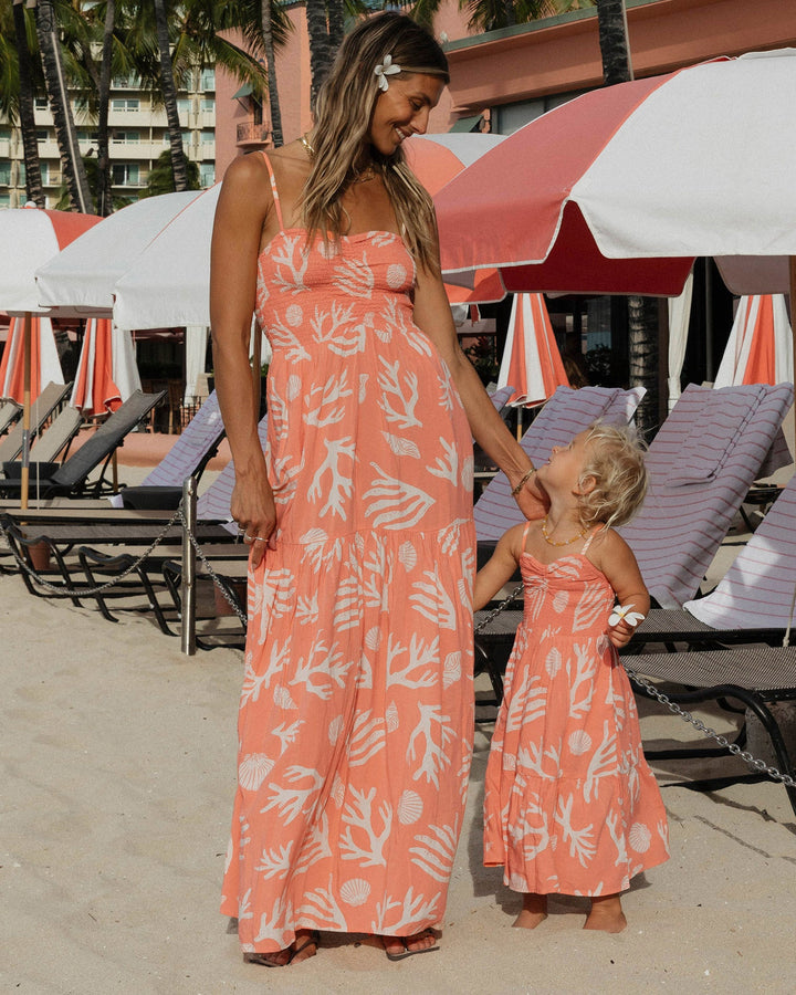 Woman and child in matching coral dresses on a beach with umbrellas and lounge chairs.