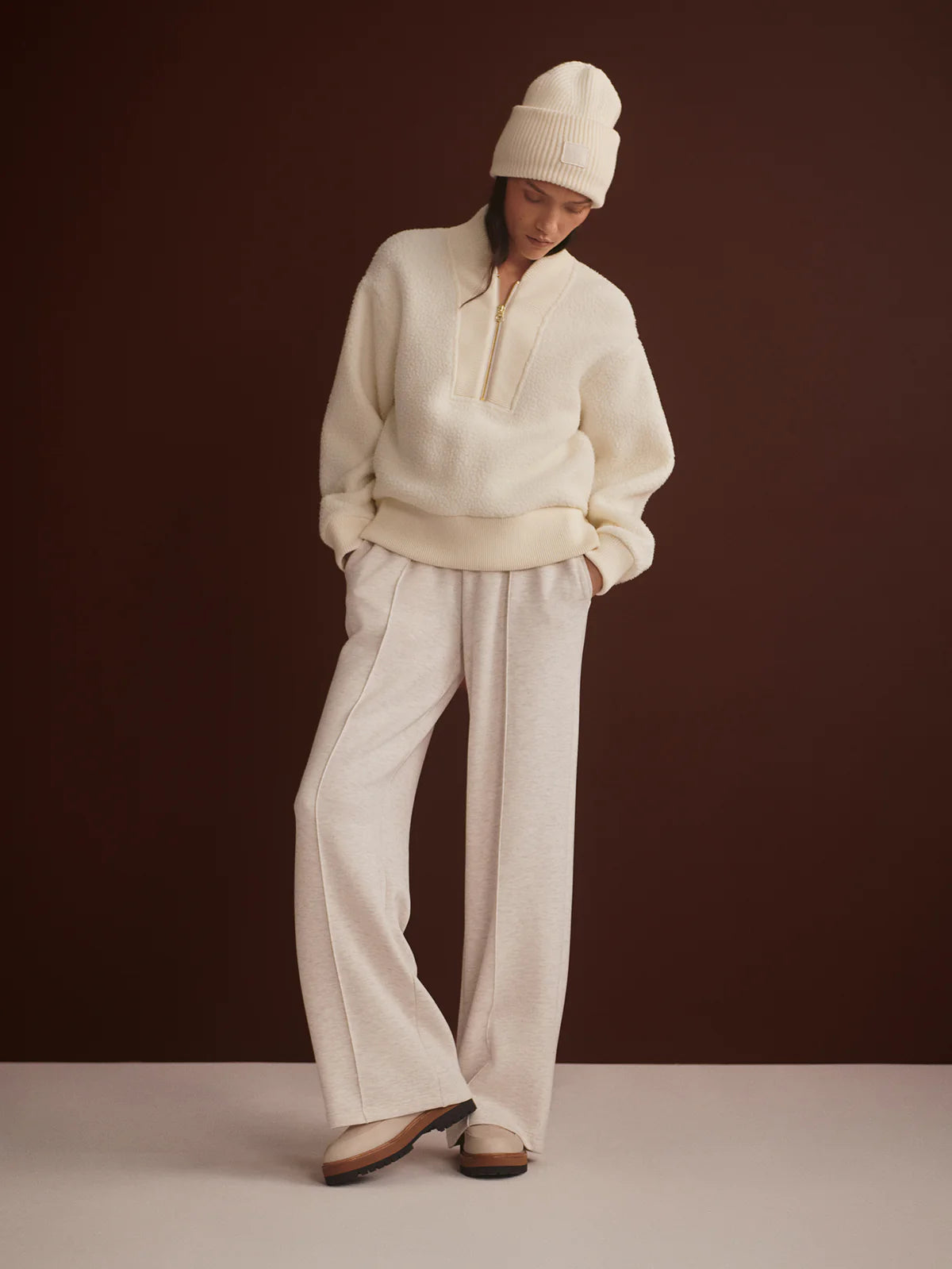 Person wearing a cream-colored sweater and pants with a white beanie against a dark brown background