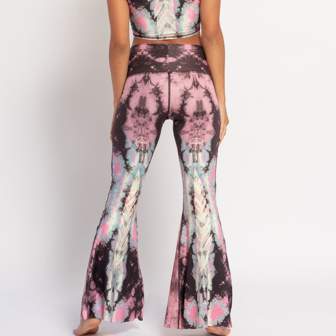 A model is shown from the back wearing crop bell bottom pants with a tie-dye-like print in pink and black tones, with native American eagle style art paired with a matching sleeveless top.